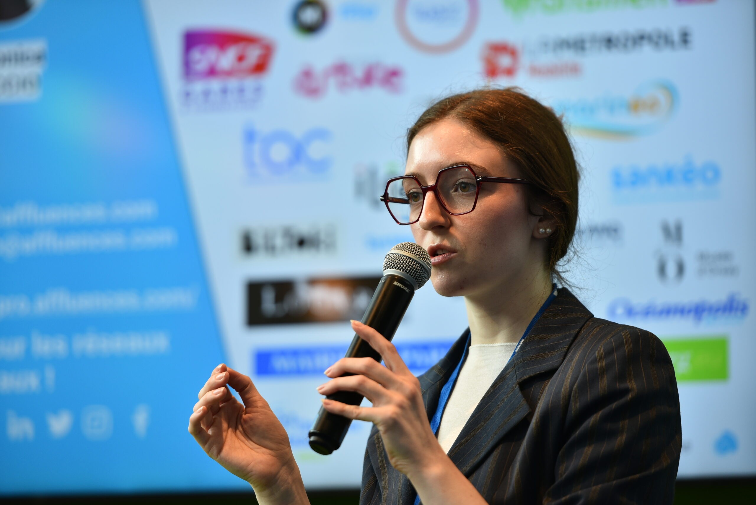 Woman speaking into a microphone in front of a screen displaying company logos