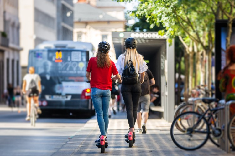 Two girls riding e-scooters near a bus stop