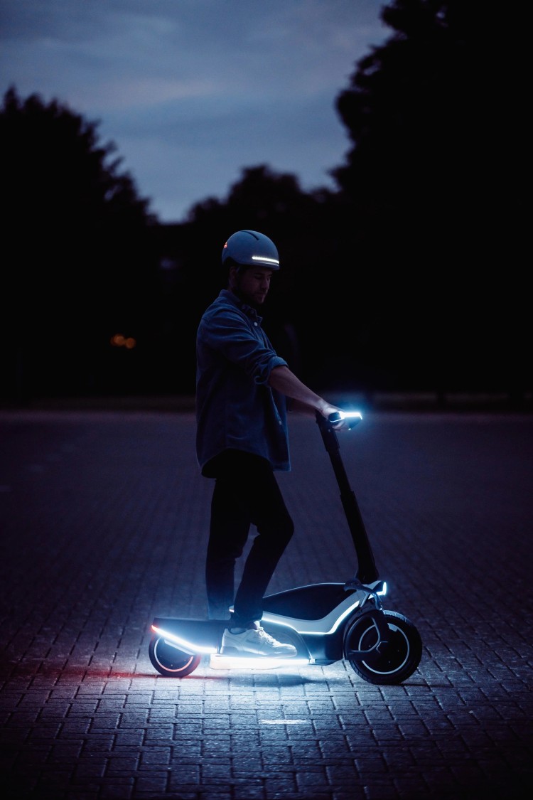 Man stands in the dark with scooter lit up at the handle bars and base by white glowing light