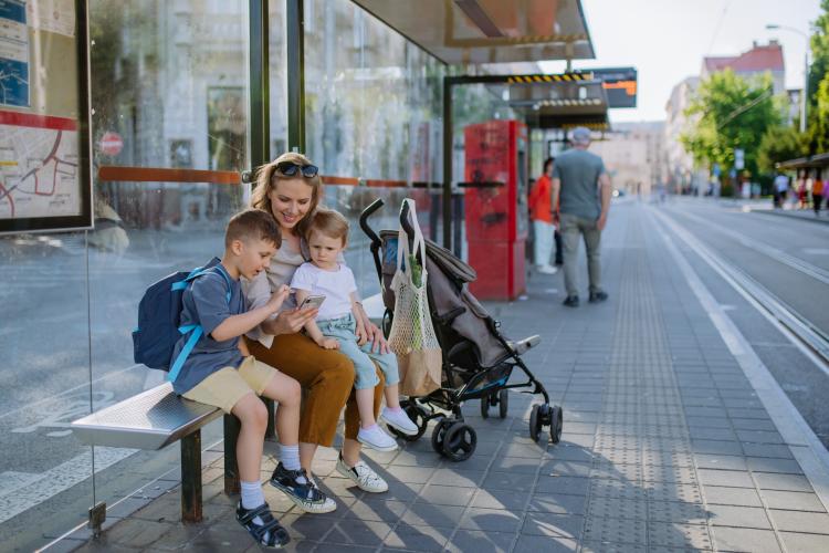 Young mother with little kids waiting on bus stop in city, scrolling on mobile phone.