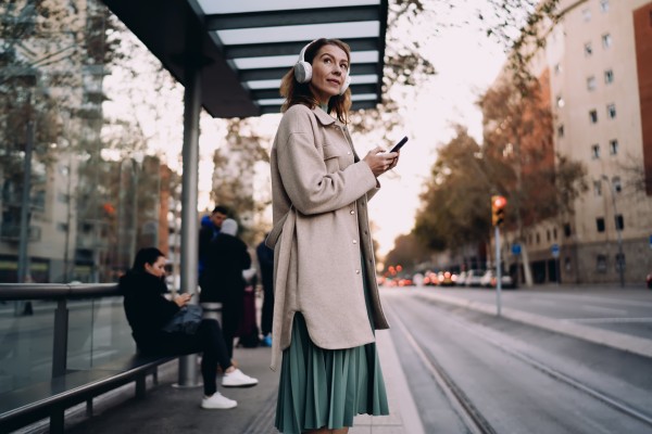 woman waiting at urban bus stop with headphones, stylish lady using smartphone app