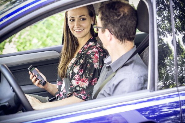 A woman in the front passenger seat of a car holds a phone with an app on screen. She is smiling at the driver in the driver's seat