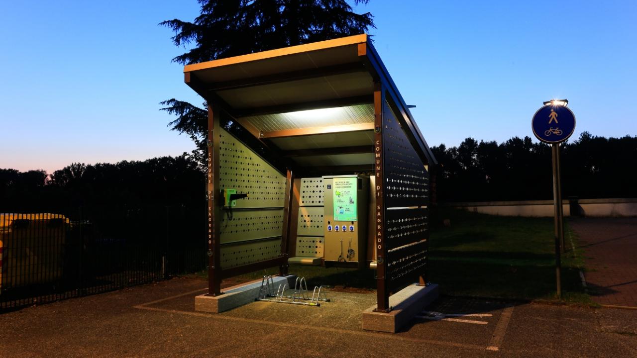 Side view of a solar-powered shelter with perforated panels, placed near a park pathway.