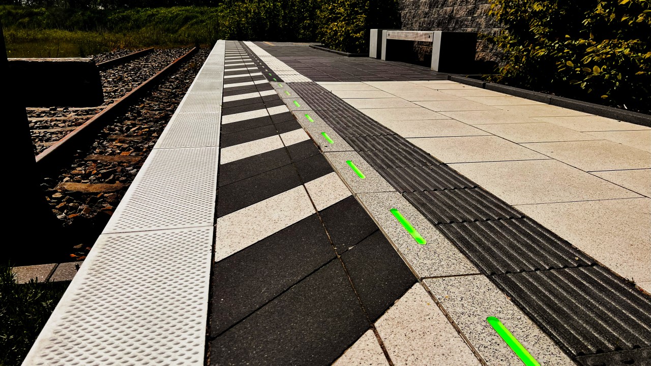 View of a train platform with green illuminated edge lights integrated into the concrete surface in outdoor daylight.