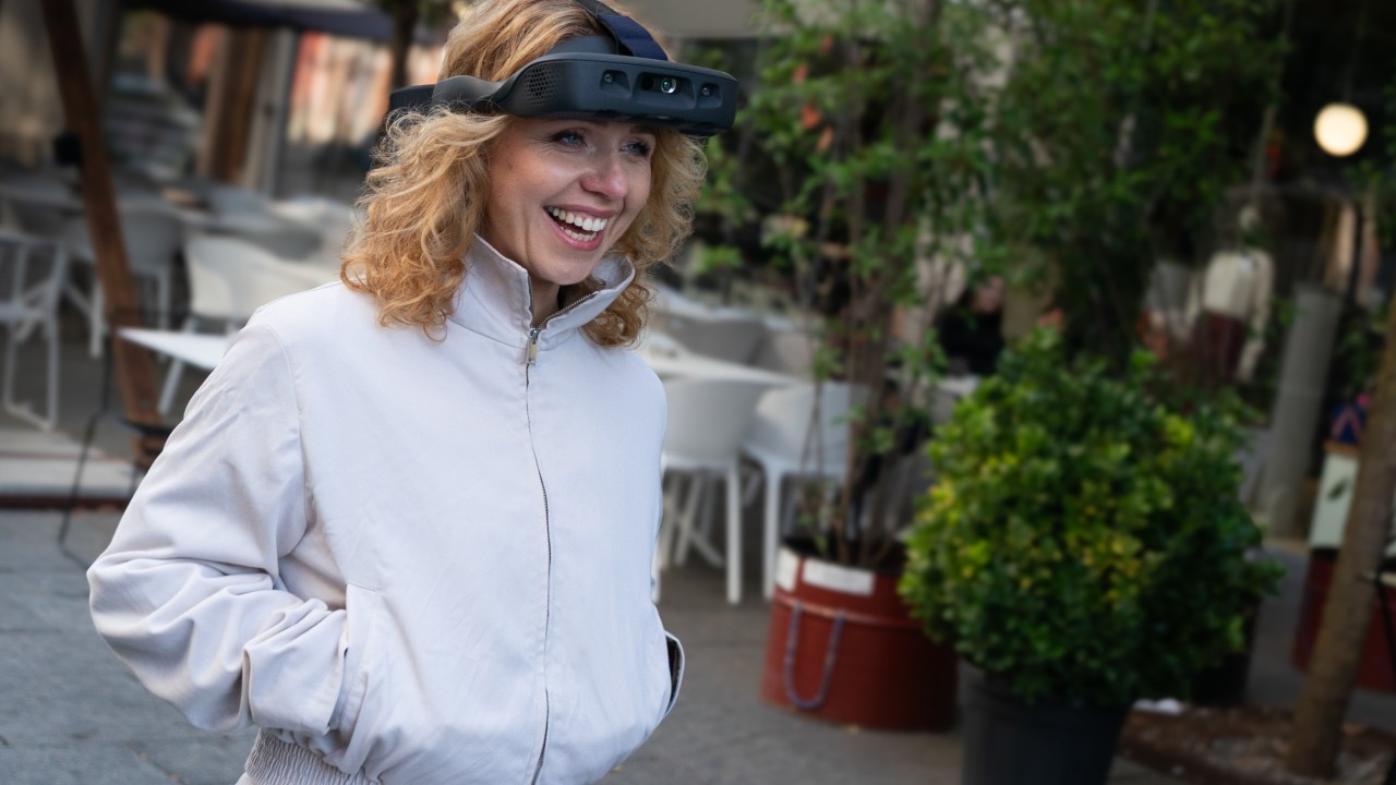 Irina Crocker smiling while wearing the .lumen Glasses for the Blind outdoors on a pedestrian street. The AI-powered wearable is clearly visible as she stands in front of cafés and greenery in an urban setting.