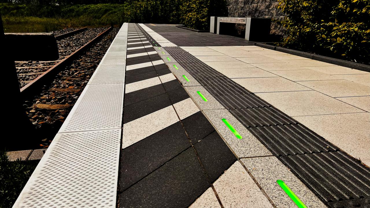 View of a train platform with green illuminated edge lights integrated into the concrete surface in outdoor daylight.